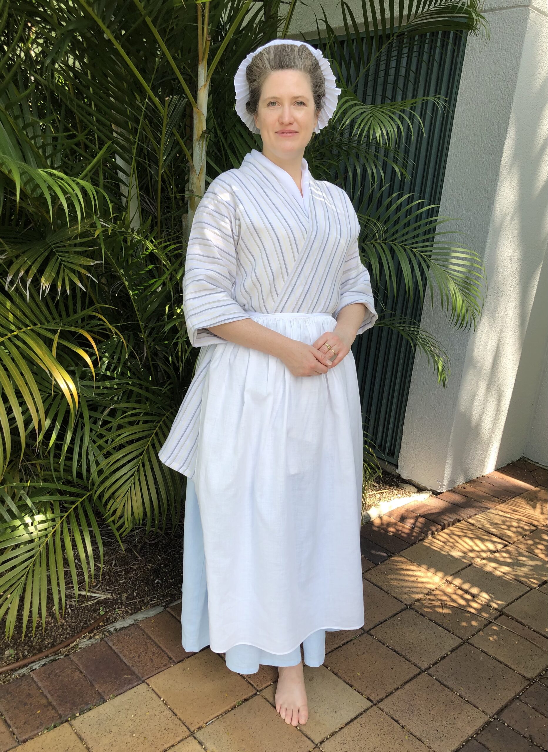 A woman smiles gently at the camera. She is wearing an 18th century working class outfit of striped linen bedgown, sky blue linen petticoat and a white linen apron.