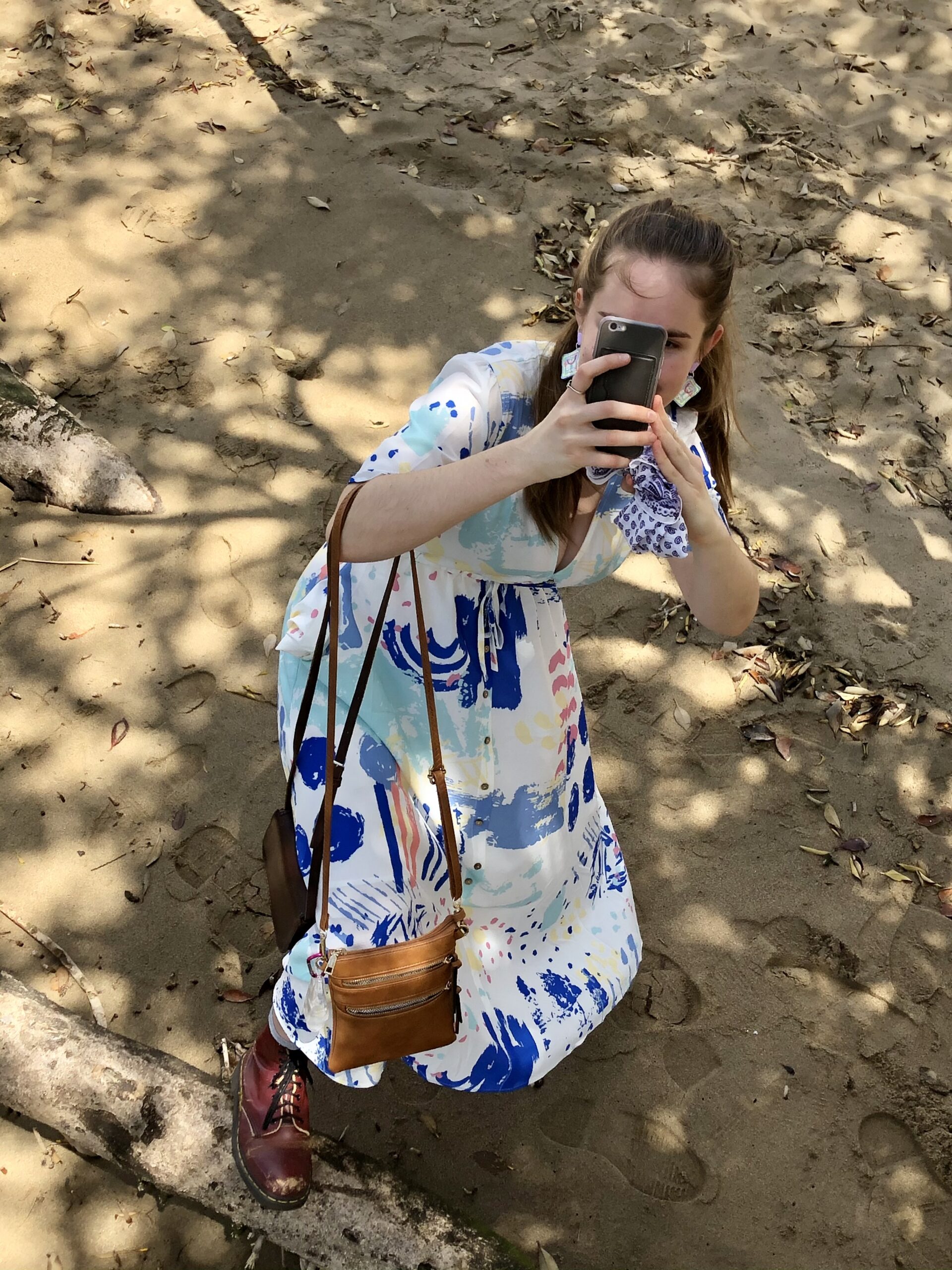 The camera looks down on a Earandthere, a woman in a white maxi dress splashed with pastel paint strokes. She is looking back at the viewer through a cell phone camera