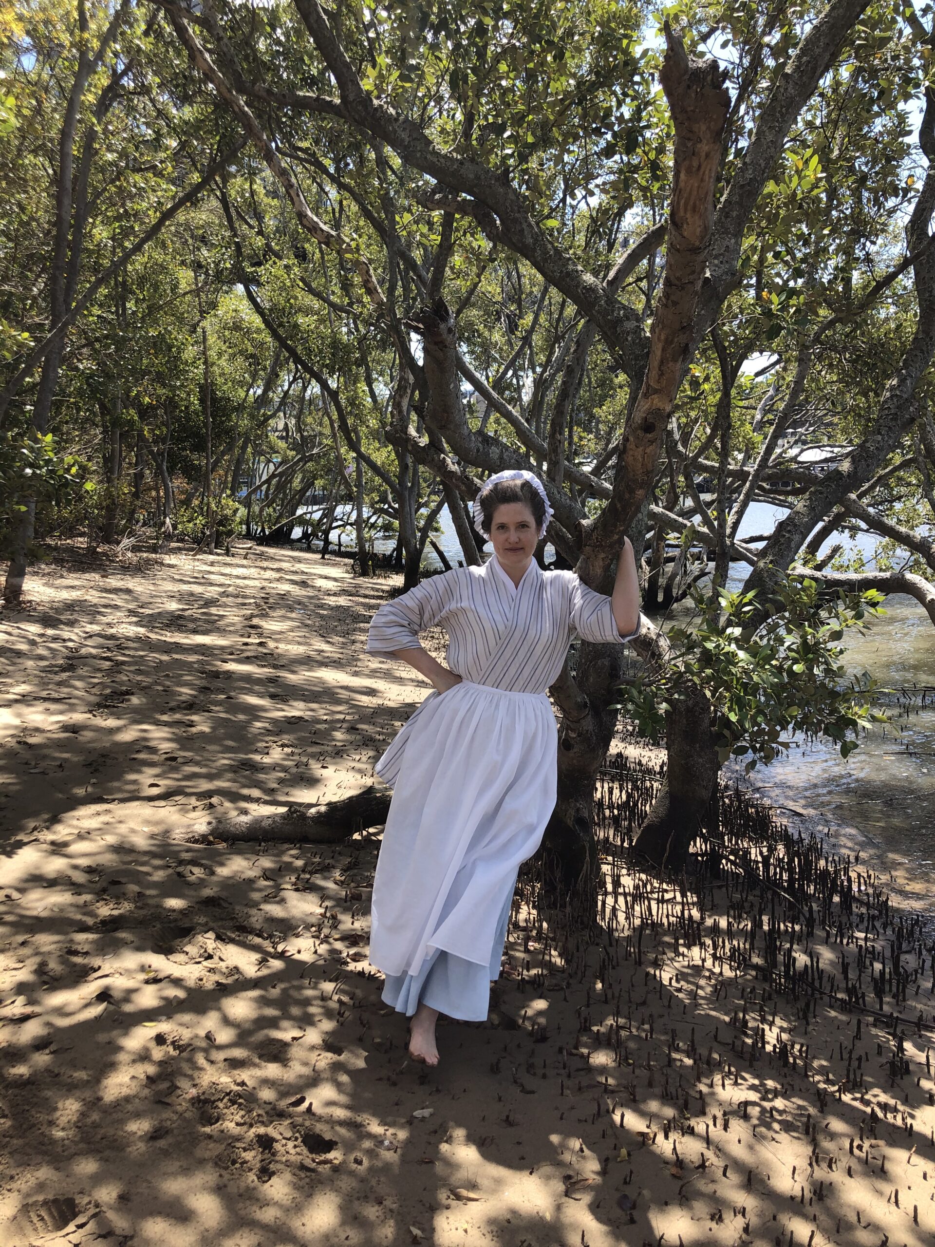 A woman leans against the trunk of a  mangrove tree, holding onto the trunk with her left hand. She is wearing an 18th Century bedgown, petticoat and apron. She is smiling.