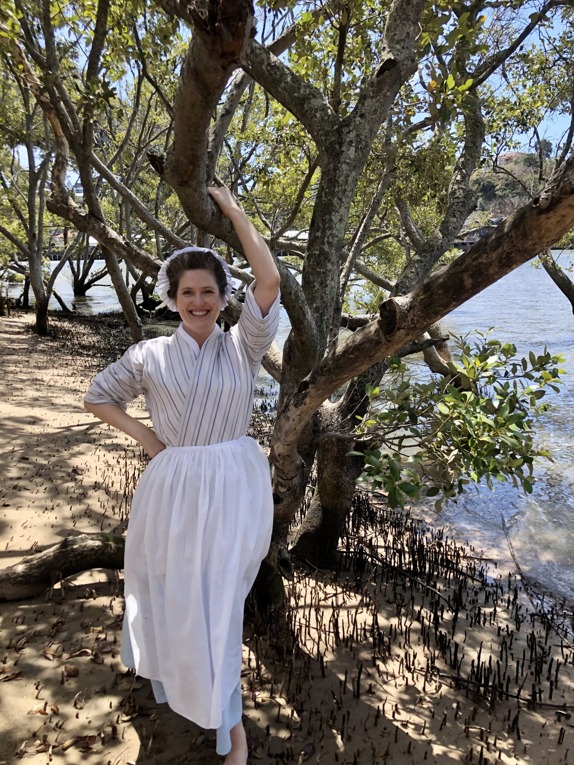 A smiling woman hangs off of a big branch of a mangrove tree. There is water beside her but under the tree the light is dappled and soft. She is wearing an 18th Century bedgown, petticoat and apron. She is smiling.