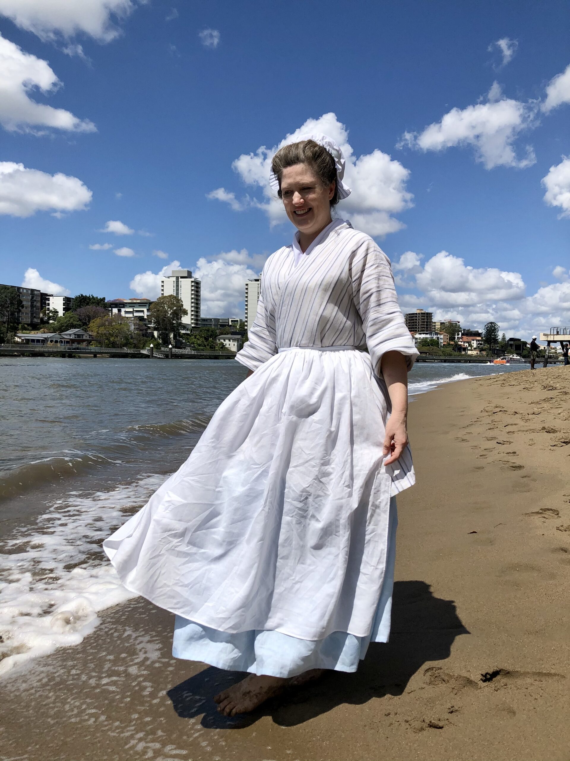 A woman wearing an 18th Century bedgown, apron and petticoat strides down the beach of the Brisbane River. The water makes small waves on the sand and her skirts and cap billow like flags in the wind