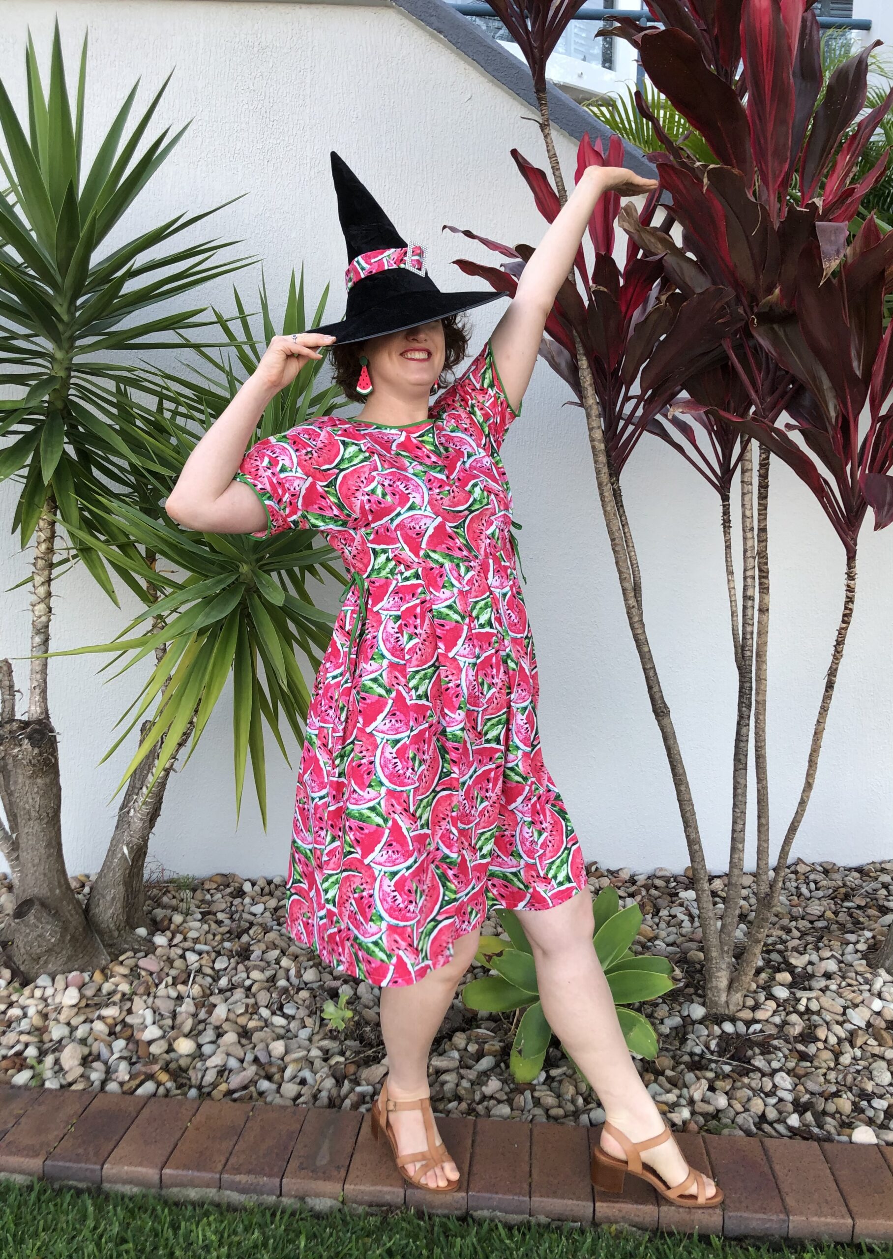 A woman strikes a pose wearing a witch hat with a pink band and a knee length dress sewn from pink and green watermelon print cotton. She would be elegant, except that her hat has fallen down over her eye.