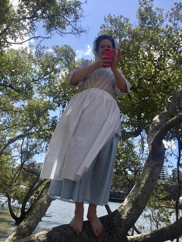 A woman stands in a mangrove tree, looking down at the viewer through the lens of a cell phone camera. She is wearing an 18th Century bedgown, petticoat and apron. 
