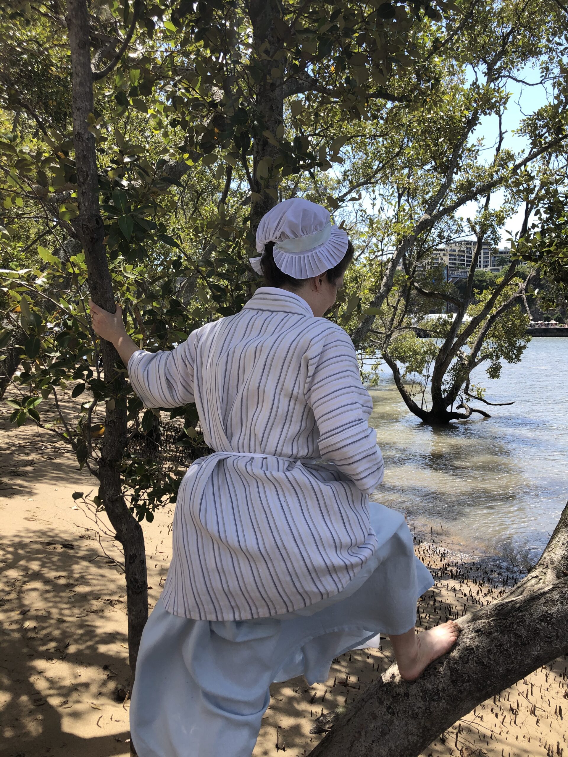 A woman is hiking up her blue skirts and climbing a mangrove tree. We are looking at the back of her - she is wearing a striped linen bedgown, a blue linen petticoat and a small white cap.