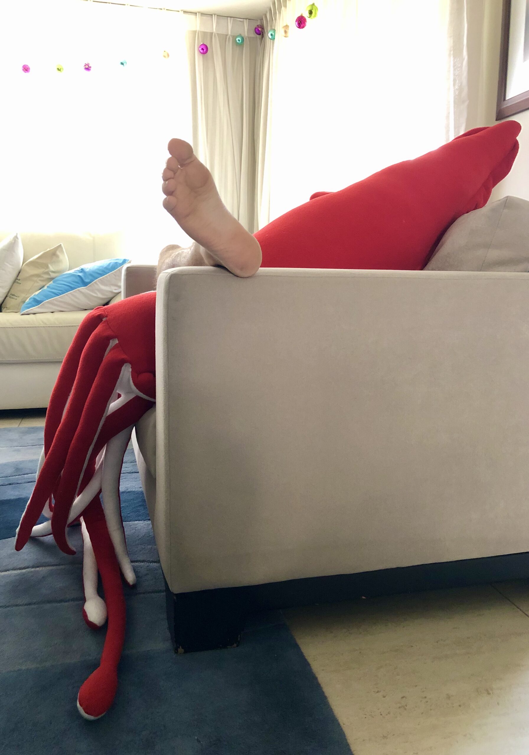 A human foot peers over the end of a grey sofa.  A giant red squid - mantle and tentacles, appears to take up the rest of the sofa.