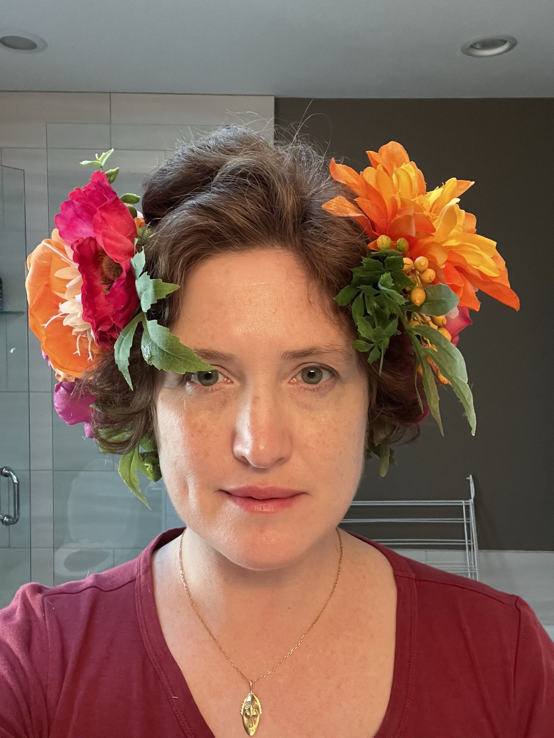 A woman stares directly in the camera. She is wearing a burgundy tshirt and on her head is an alphonse mucha inspired headband heavy with flowers.