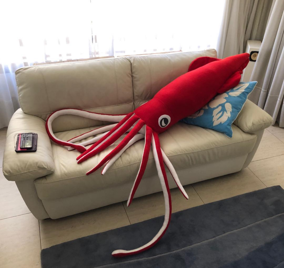 A giant stuffed Christmas squid spreads its tentacles across a white leather sofa. It is the same size as the sofa.